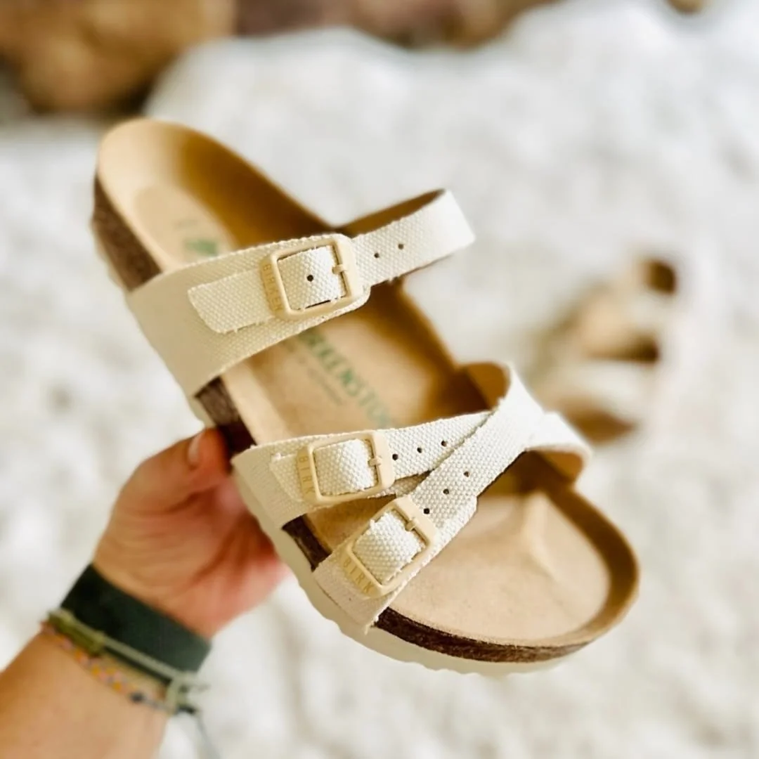 Cream-colored Birkenstock Franca sandal with three canvas straps and tonal buckles, featuring a cork footbed and white EVA sole, held by a hand against a soft, blurred background.