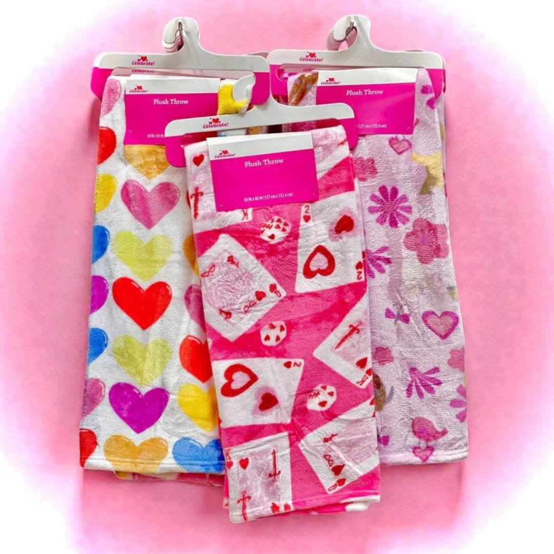 Celebrate! Valentine's Day plush throw blankets in vibrant patterns, including colorful hearts, playing card designs, and floral motifs, displayed on white hangers against a pink background.