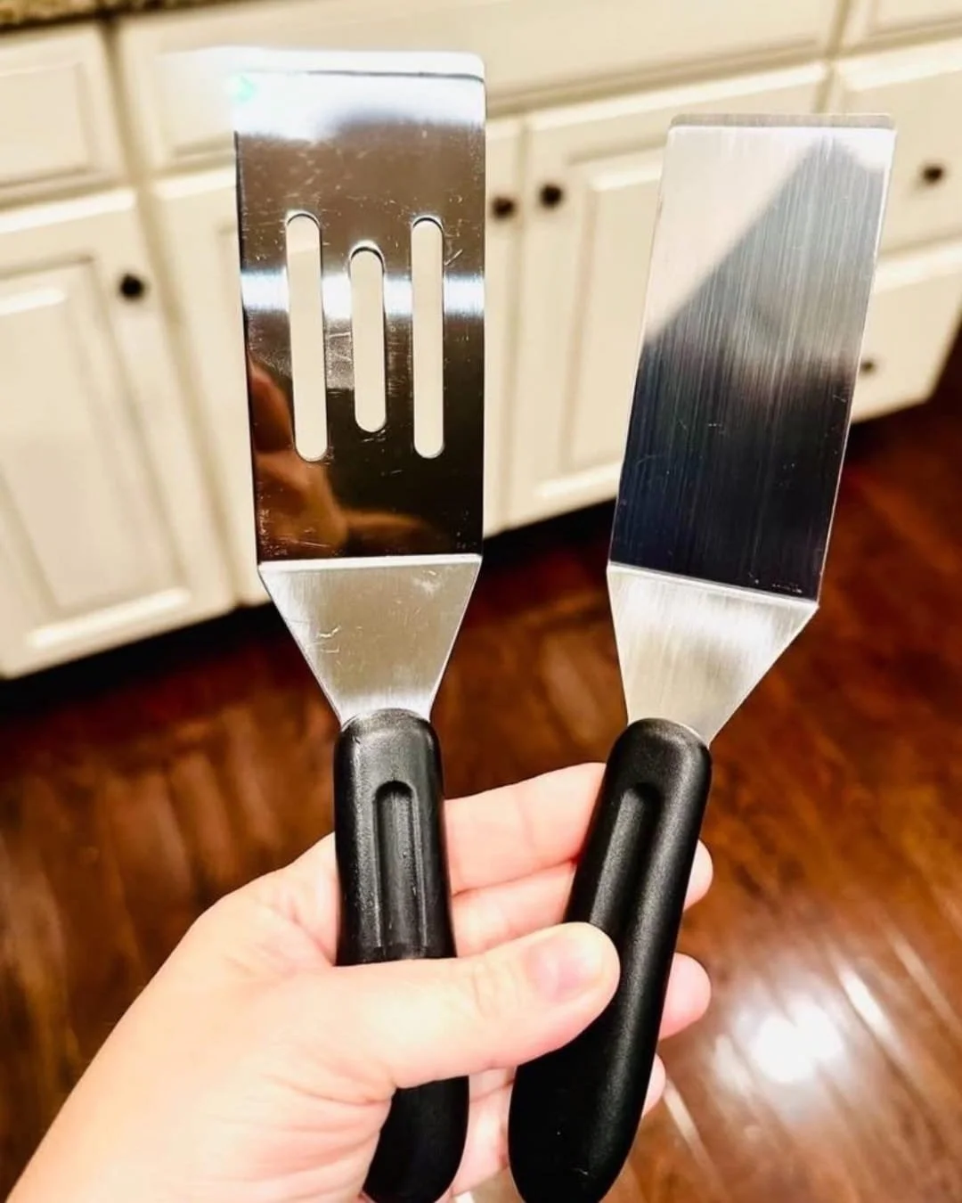Close-up of a hand holding two stainless steel spatulas, one slotted and one solid, in a modern kitchen with white cabinets and dark wood flooring in the background.