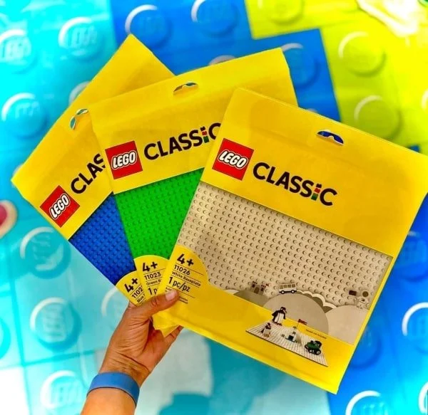 Hand holding three LEGO Classic baseplates in yellow packaging: white, green, and blue, with LEGO branding.