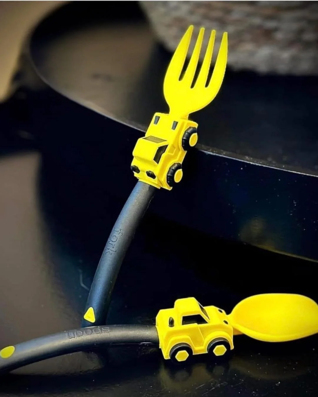 Children's construction-themed utensils featuring a yellow fork and spoon with toy truck accents and ergonomic black handles, displayed on a dark surface with a blurred background.