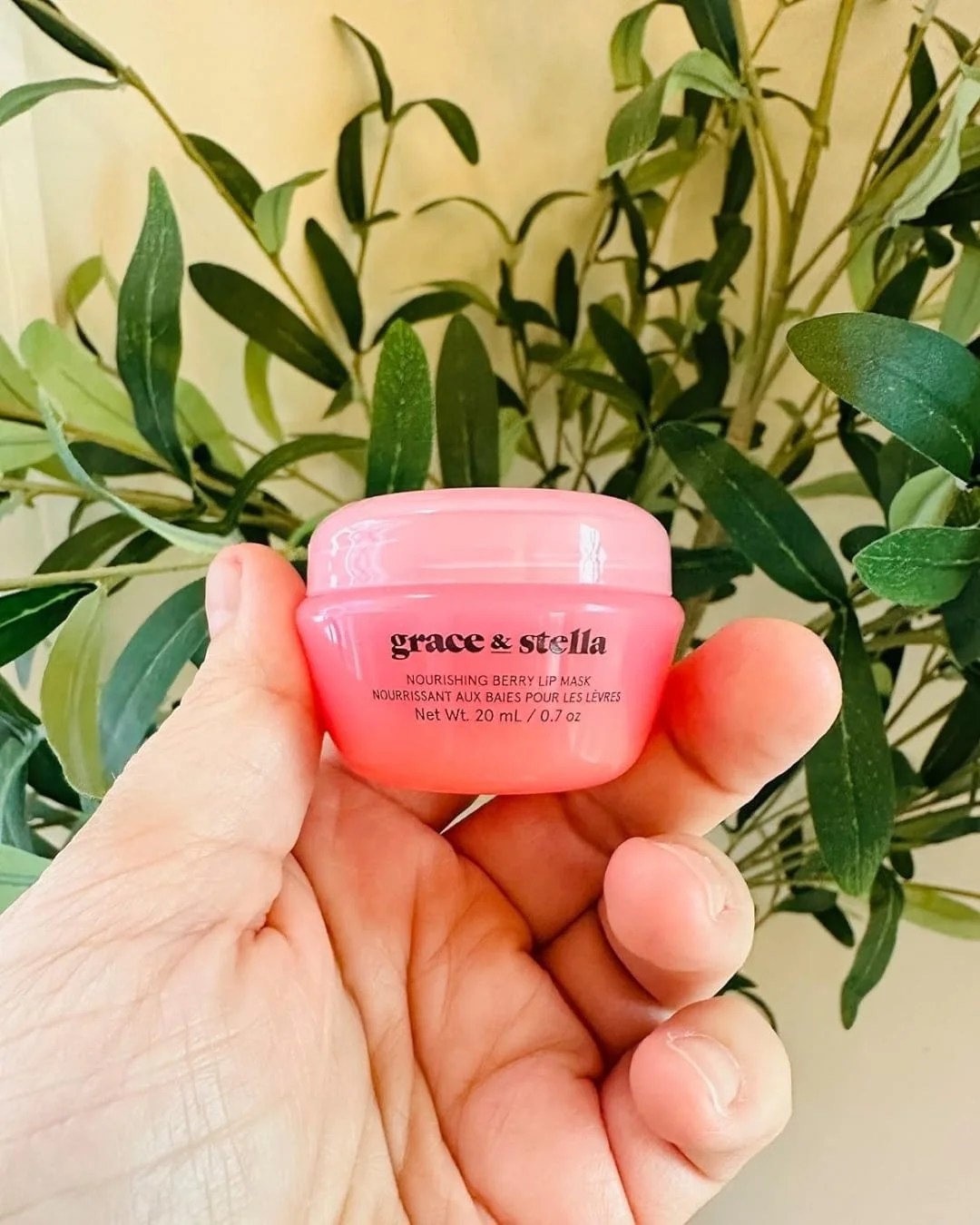 Close-up of a pink jar of grace & stella Nourishing Berry Lip Mask held in a hand, with a green leafy plant and cream-colored wall in the background.