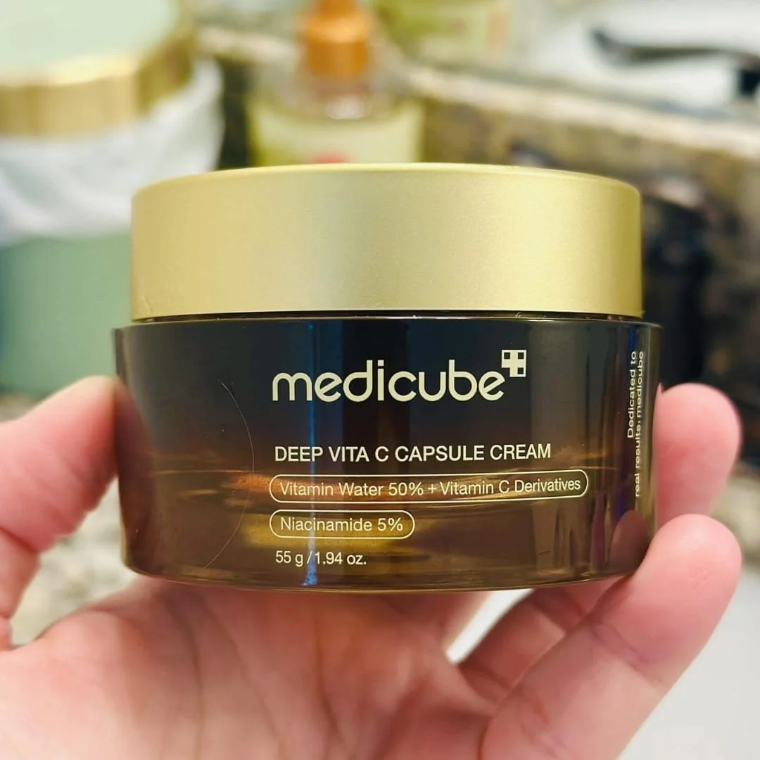 Medicube Deep Vita C Capsule Cream jar with gold lid held in hand in bathroom setting.