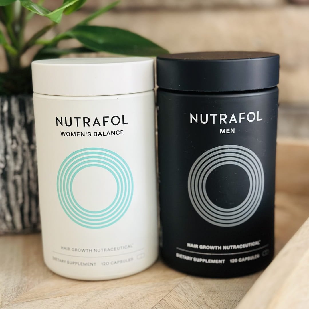 Nutrafol Hair Growth Nutraceutical supplements for Women’s Balance and Men, displayed side-by-side on a wooden surface with a green potted plant and a neutral brick-textured background.