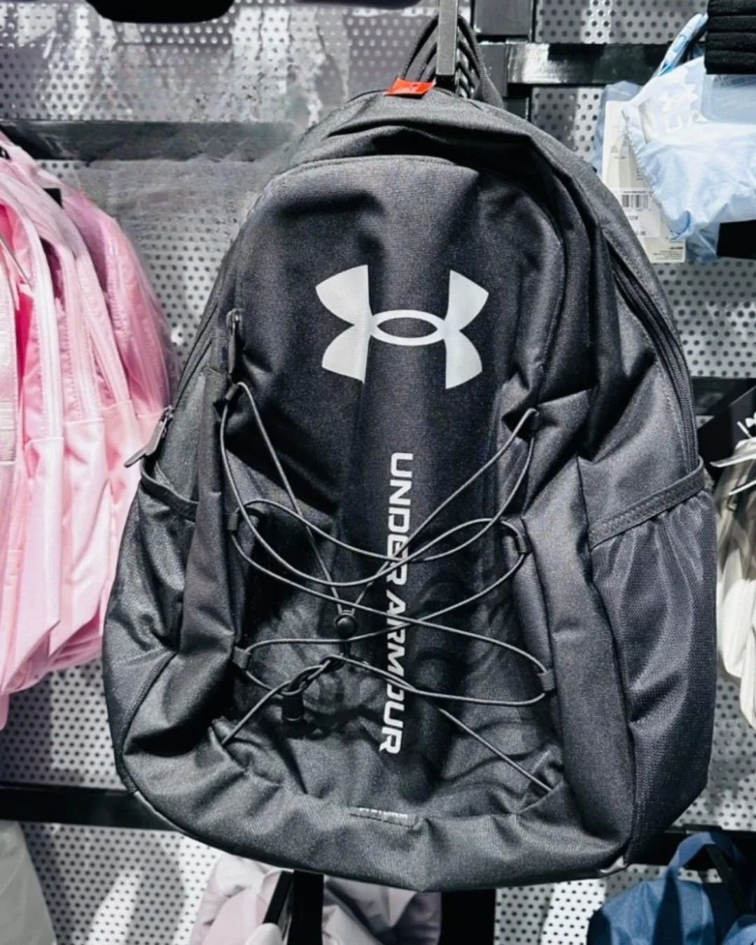 Black Under Armour backpack with white logo and vertical branding, featuring a bungee cord system and multiple compartments, displayed on a retail pegboard wall surrounded by other bags.