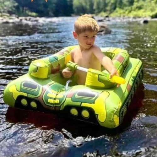 Young boy sitting in a green camouflage inflatable tank float in a body of water, holding a yellow and green water squirter, with a forested shoreline in the background.
