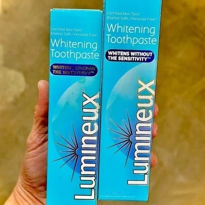 Two blue Lumineux Whitening Toothpaste boxes, highlighting Non-Toxic, Peroxide Free, and Whitens Without Sensitivity, held against a beige background.