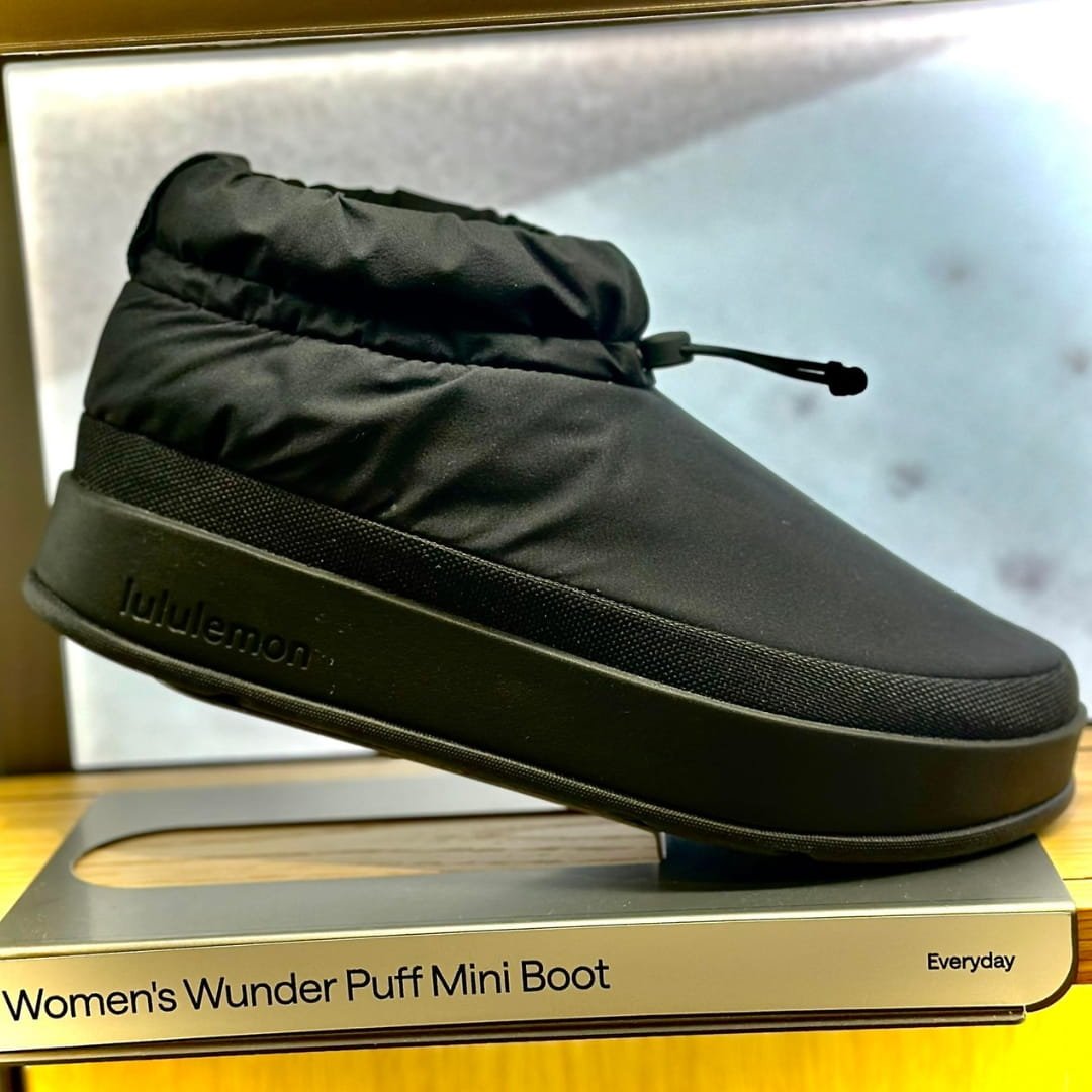 Lululemon Women's Wunder Puff Mini Boot in black with a quilted upper, drawstring closure, and thick sole featuring embossed branding, displayed on a metallic retail stand.