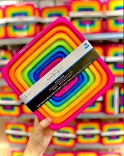 Hand holding a 14-piece Rainbow Food Storage Set with nested containers in vibrant colors from pink to blue, against a backdrop of shelves filled with similar colorful containers.