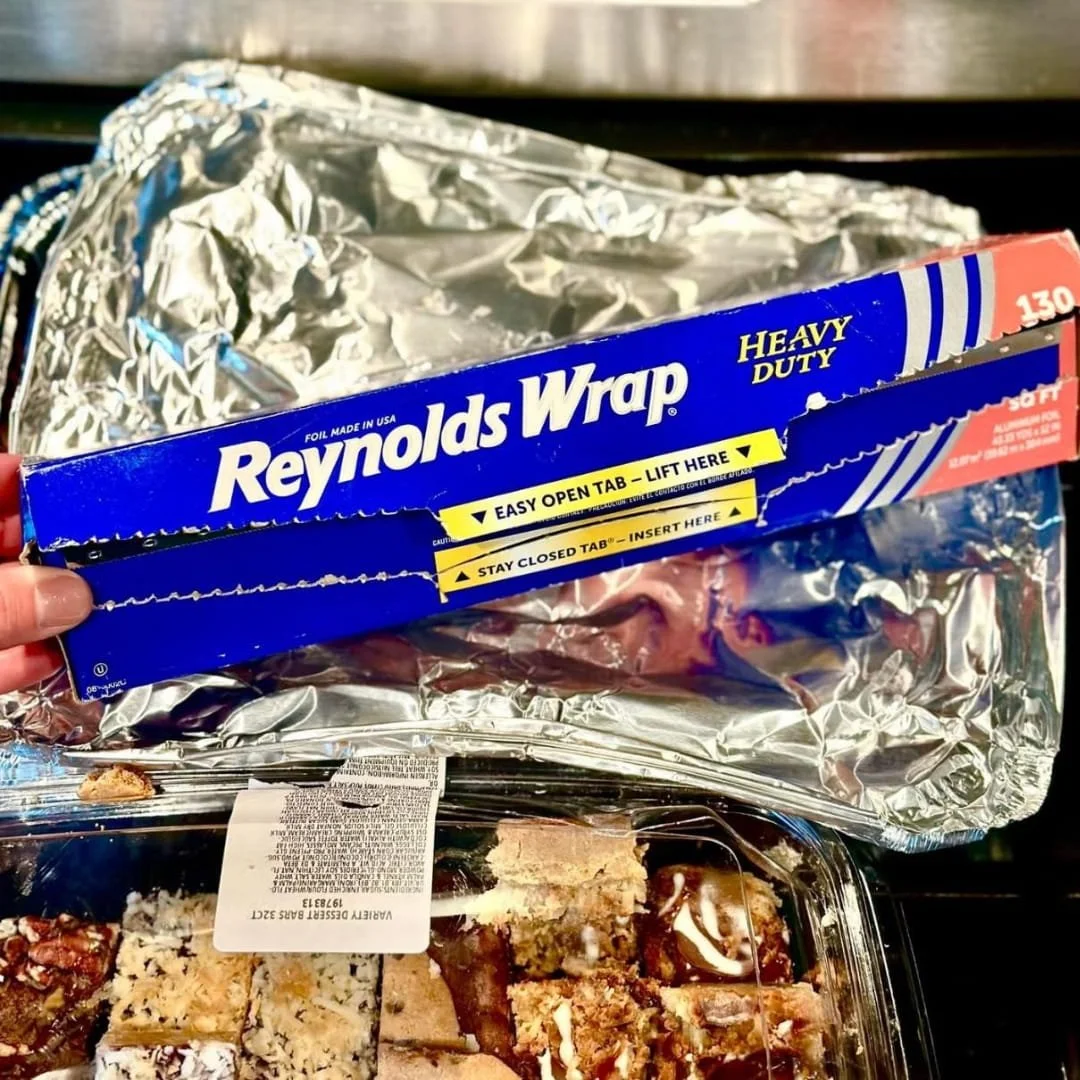Reynolds Wrap Heavy Duty aluminum foil box held above a container of assorted dessert bars, with crumpled foil in the background, showcasing practical food storage use.