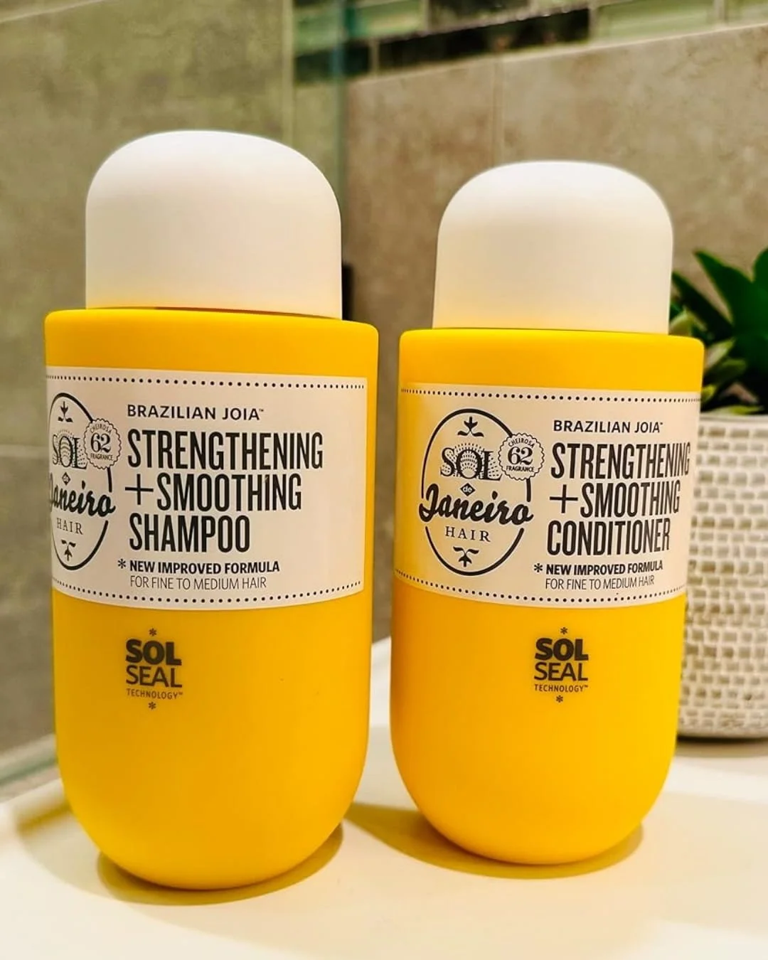 Sol de Janeiro Brazilian Joia Shampoo and Conditioner set in bright yellow bottles with white caps, displayed on a light surface with a plant and tiled background.