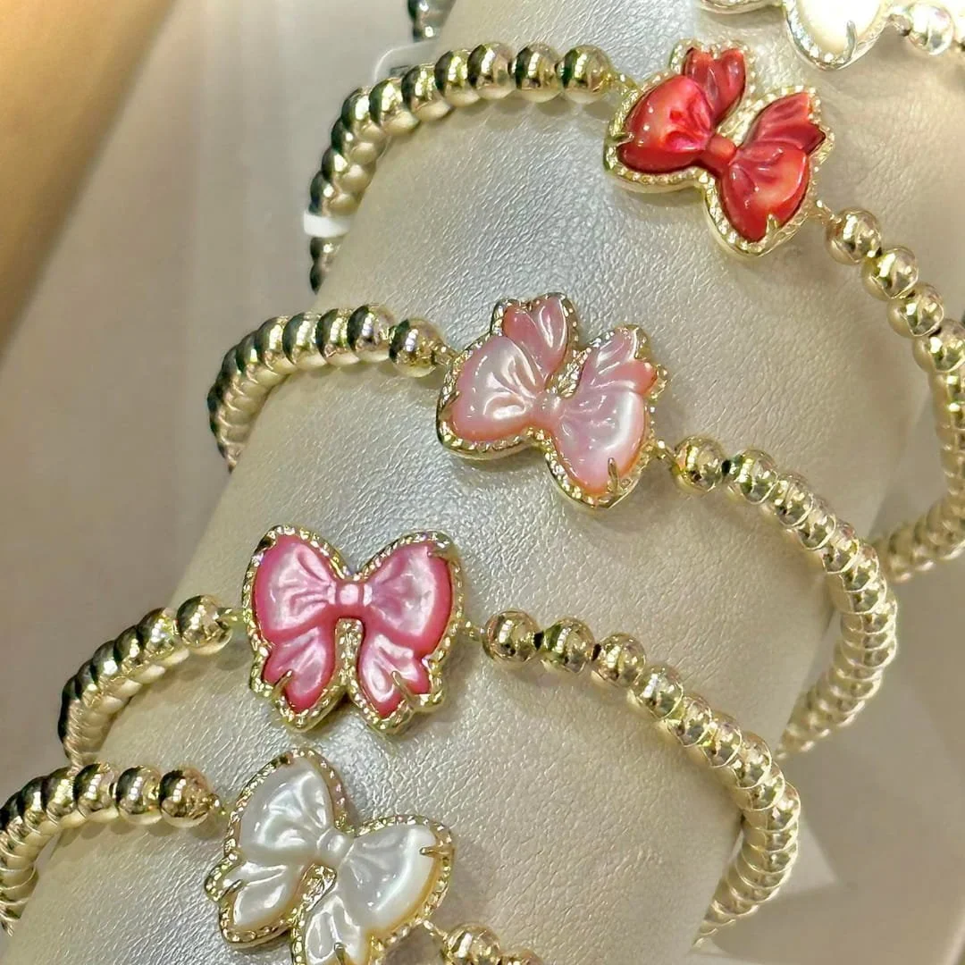 Kendra Scott gold beaded bracelets with iridescent bow-shaped charms in red, pink, and white, displayed on a cream velvet stand.