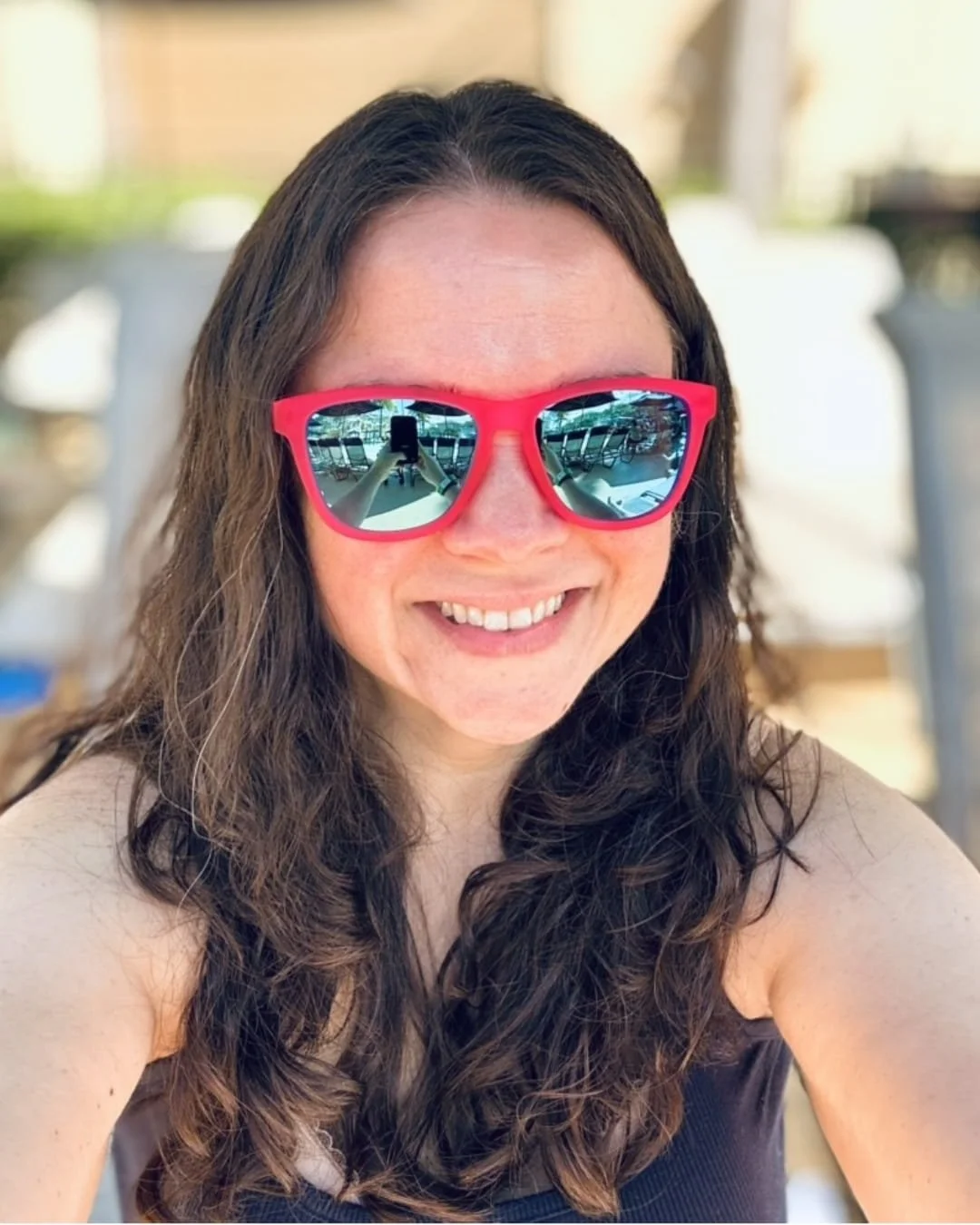 Nicole wearing bold red-framed Goodr sunglasses with mirrored blue lenses, reflecting a poolside setting with lounge chairs and umbrellas, taken in bright sunlight.