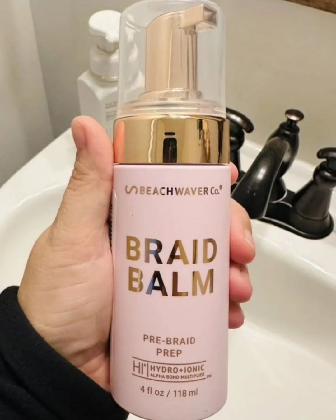 Hand holding Beachwaver Primer Foam in a pink bottle with gold pump, photographed over a bathroom sink