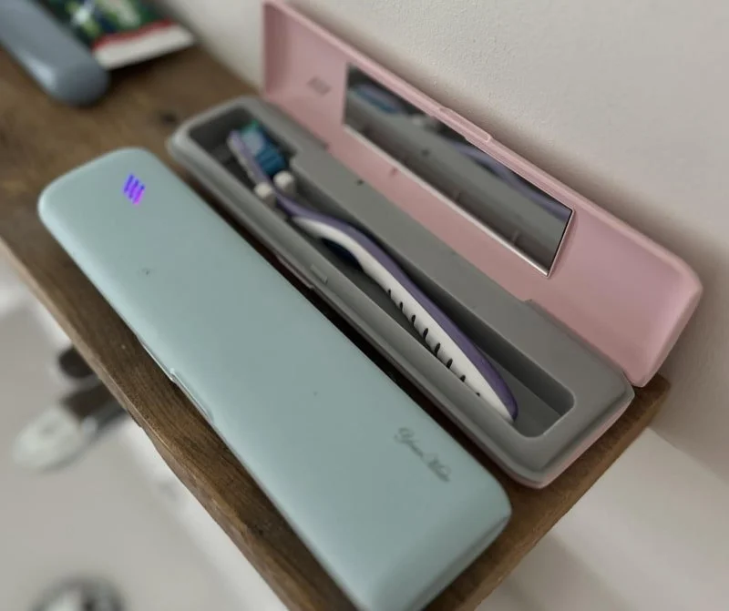 Toothbrush cases on wooden surface, featuring a light blue case with Gourmet White branding and purple indicator light, and a pink open case with a purple and white toothbrush inside, ideal for travel or UV sanitization.