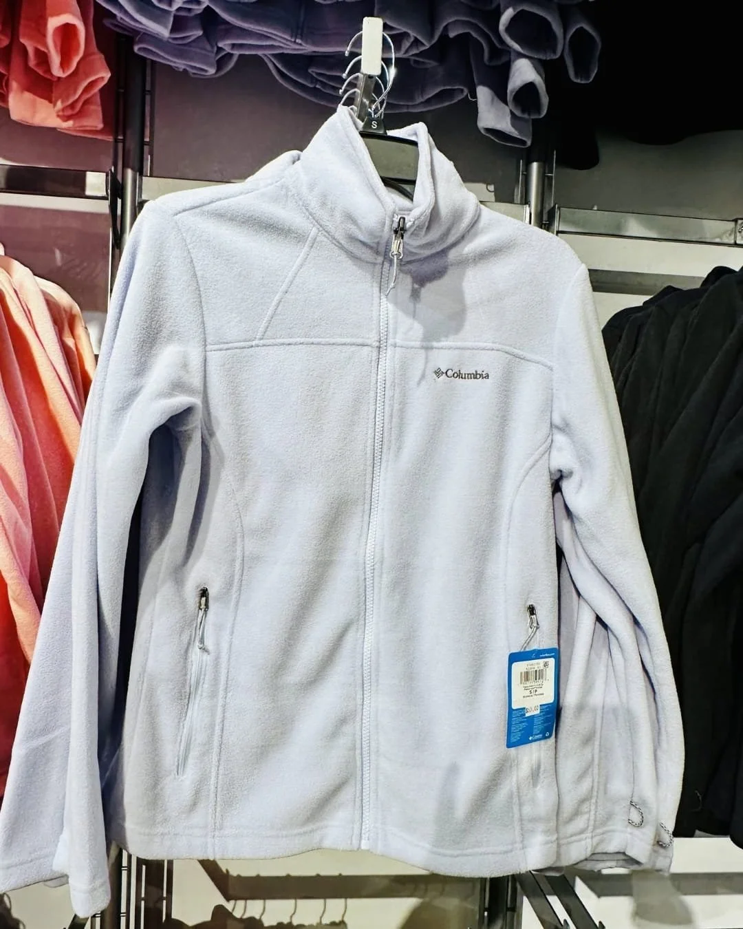 Light lavender Columbia fleece jacket with a full-zip front, zippered pockets, and high collar hanging on a retail rack with other jackets in the background