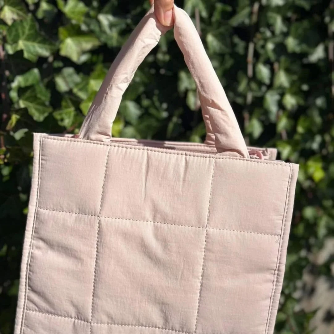 CLUCI puffer tote bag in pale pink with a quilted design, padded handles, and zipper closure, held against a vibrant green ivy background.