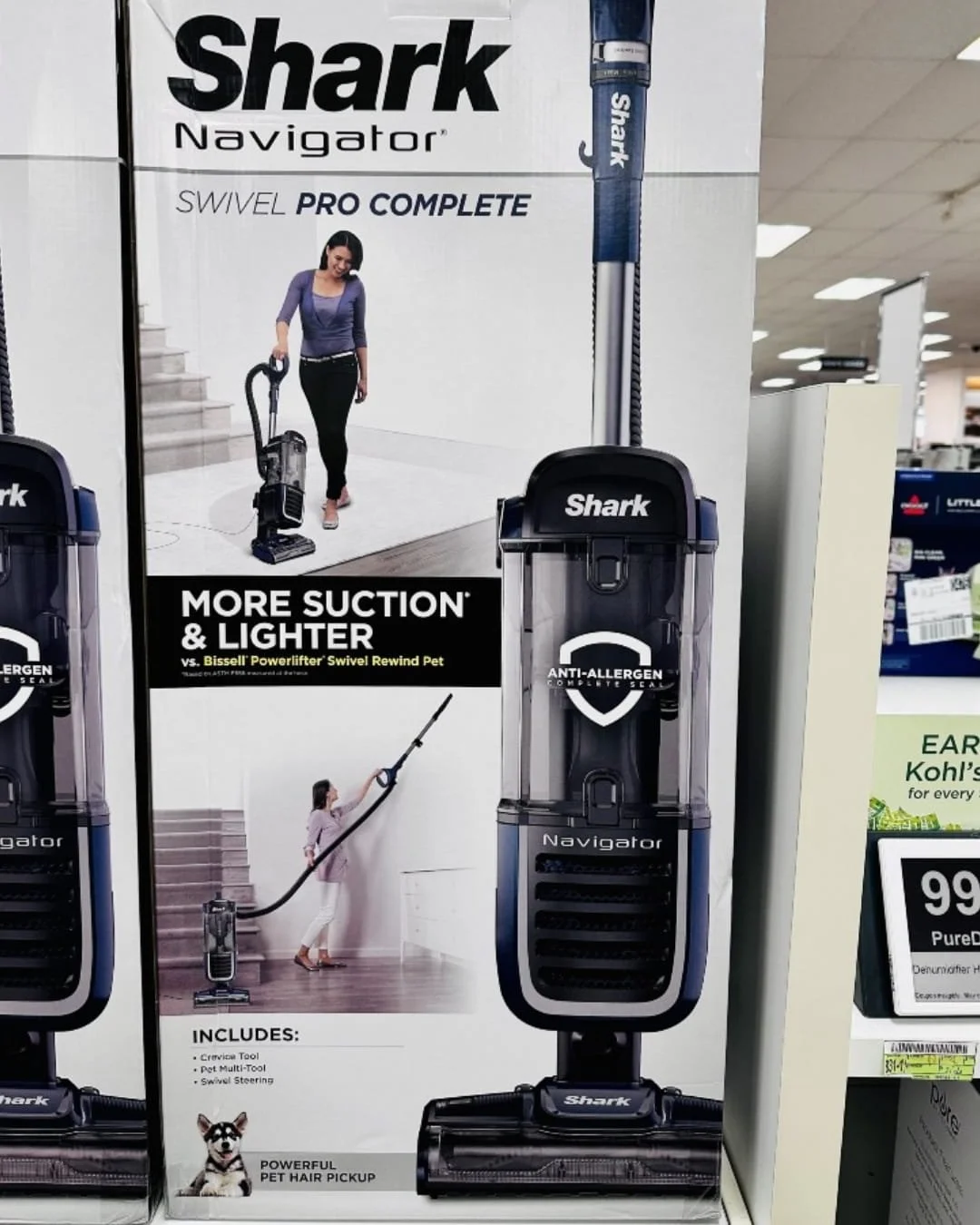 Retail box of the Shark Navigator Swivel Pro Complete upright vacuum cleaner, featuring a navy blue and silver design, with claims of more suction and lighter weight compared to competitors, and tools for pet hair pickup, displayed on a store shelf.