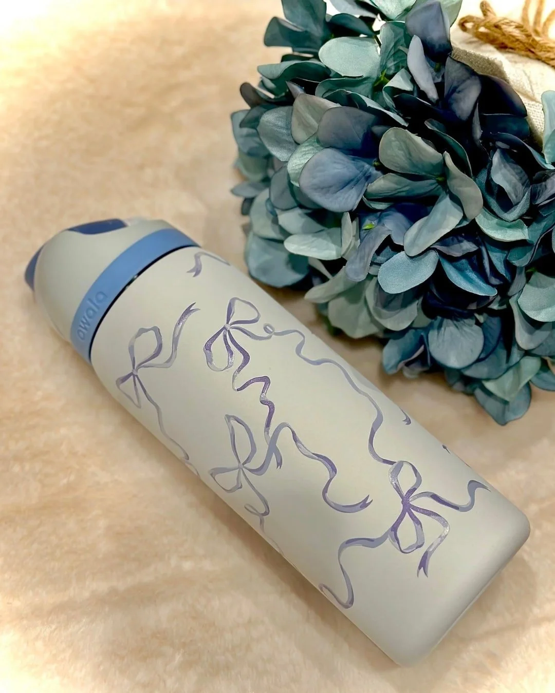 Light gray Owala water bottle with a whimsical purple ribbon and bow pattern, resting on a soft beige surface with blue-green artificial flowers in the background.