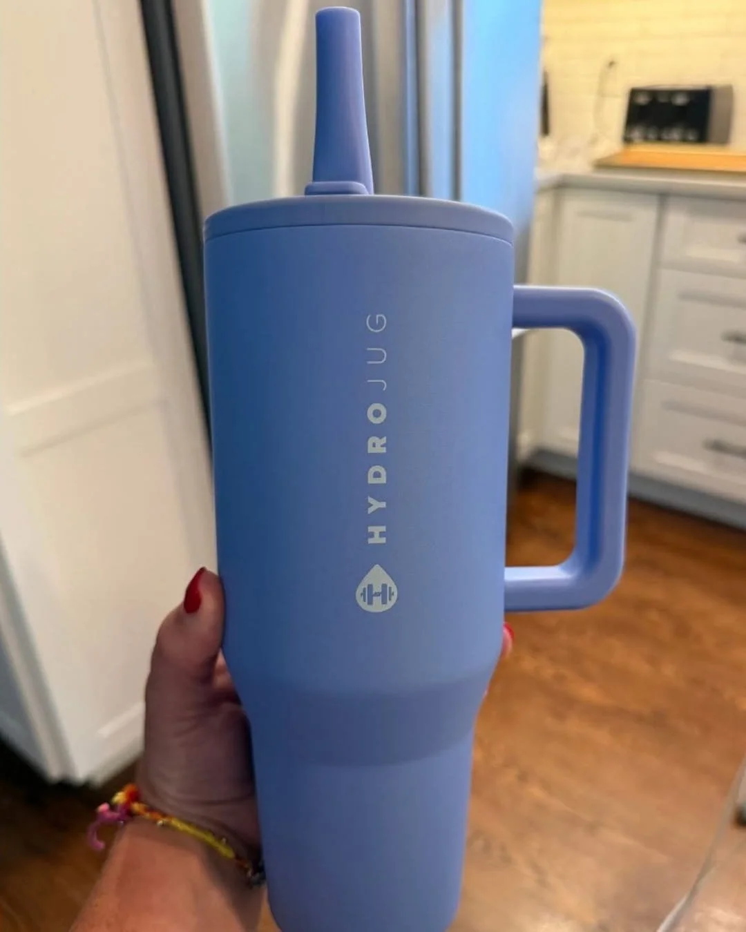 Periwinkle HydroJug water bottle with handle and straw, held indoors in front of a blurred kitchen background