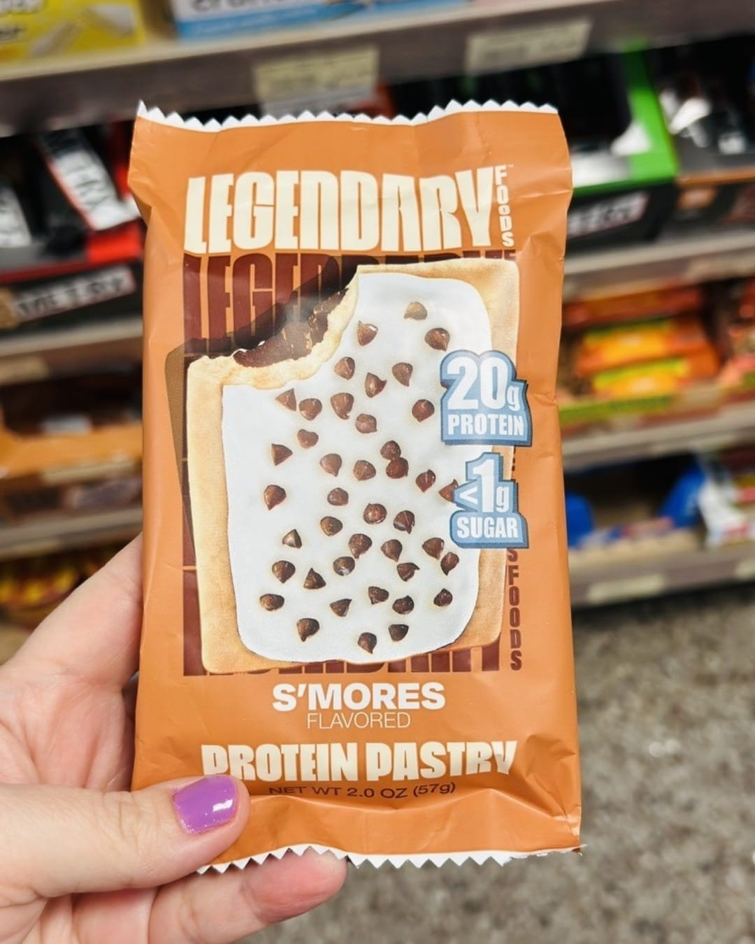 Legendary Foods S'mores Flavored Protein Pastry package held in a hand with purple nail polish, featuring 20g protein and less than 1g sugar, with an illustration of a frosted pastry and a grocery store shelf background.