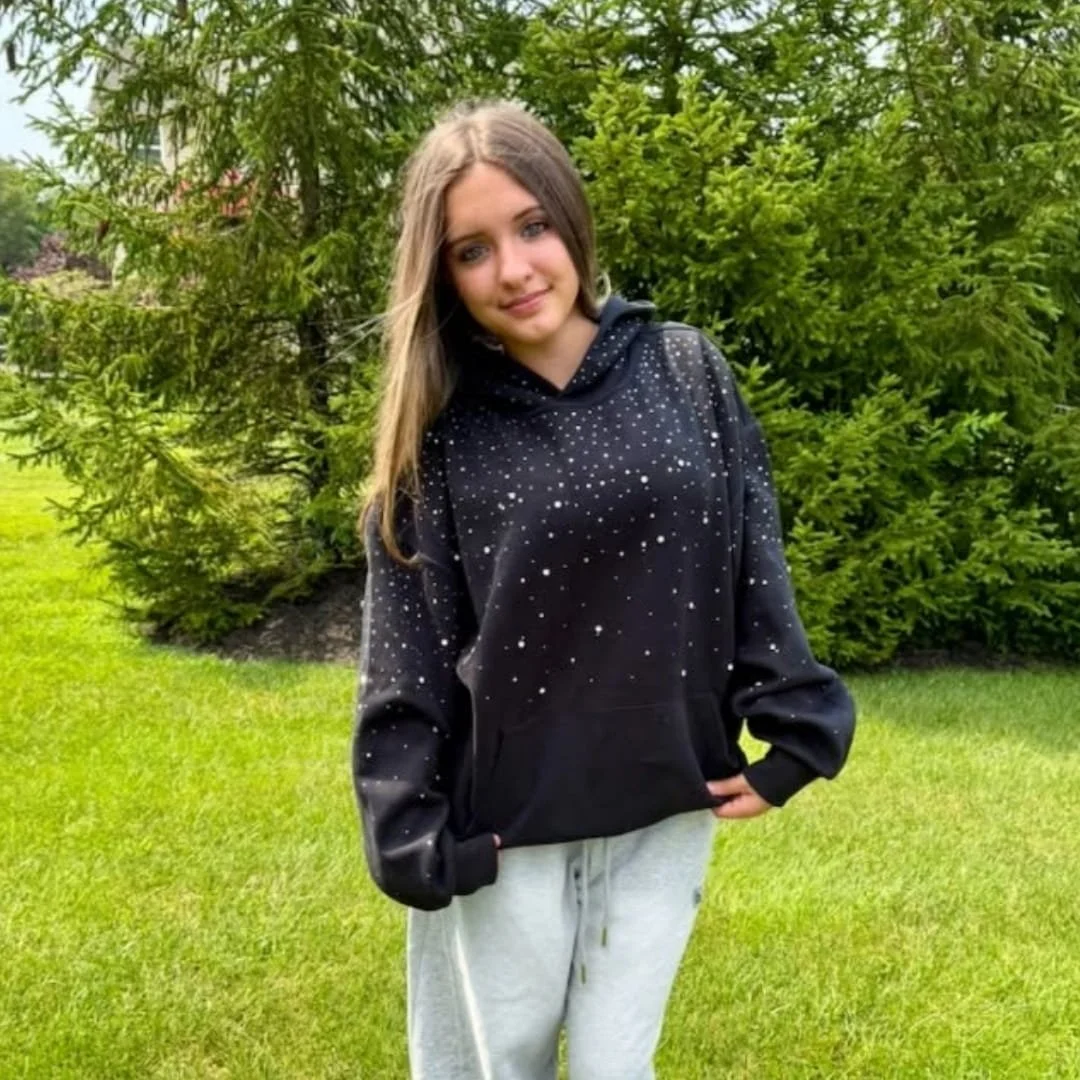 Makenzie wearing a black Aelfric Eden rhinestone hoodie with gray sweatpants while standing outdoors in front of green trees