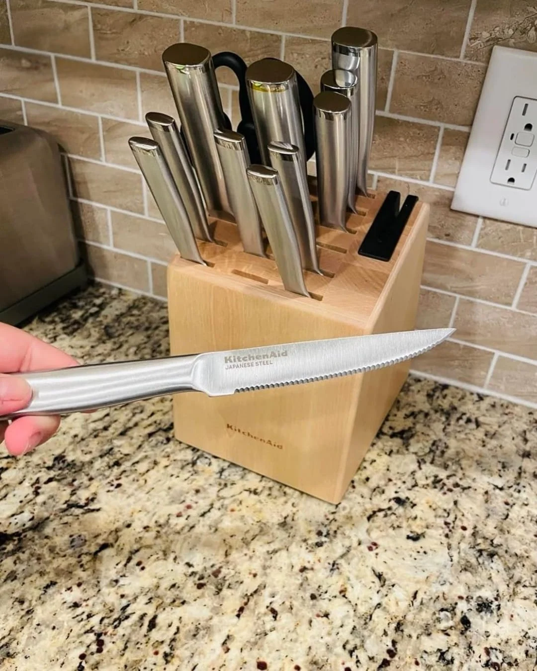KitchenAid knife set with wooden block and Japanese steel steak knife held in hand on kitchen counter.
