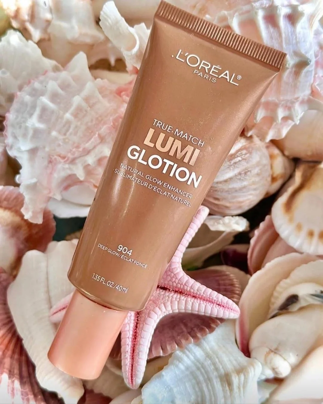 L'Oréal True Match Lumi Glotion in shade 904 Deep Glow, a natural glow enhancer, displayed on a bed of seashells and a pink starfish, evoking a beach-inspired aesthetic.