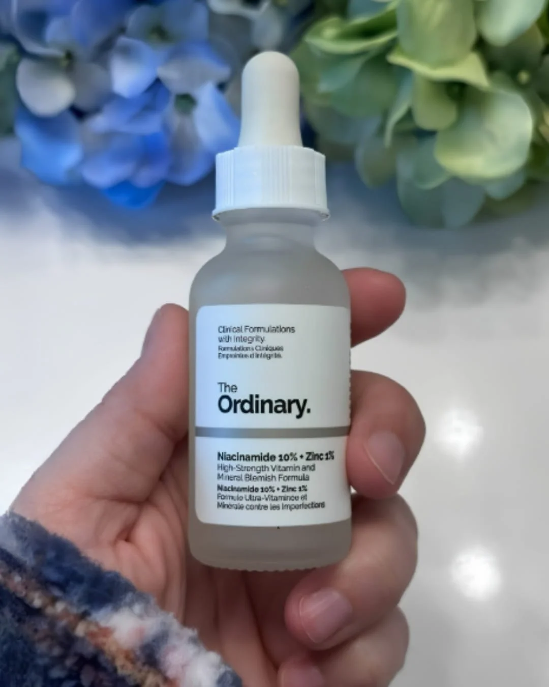 The Ordinary Niacinamide 10% Zinc 1% serum bottle held in hand with soft floral background