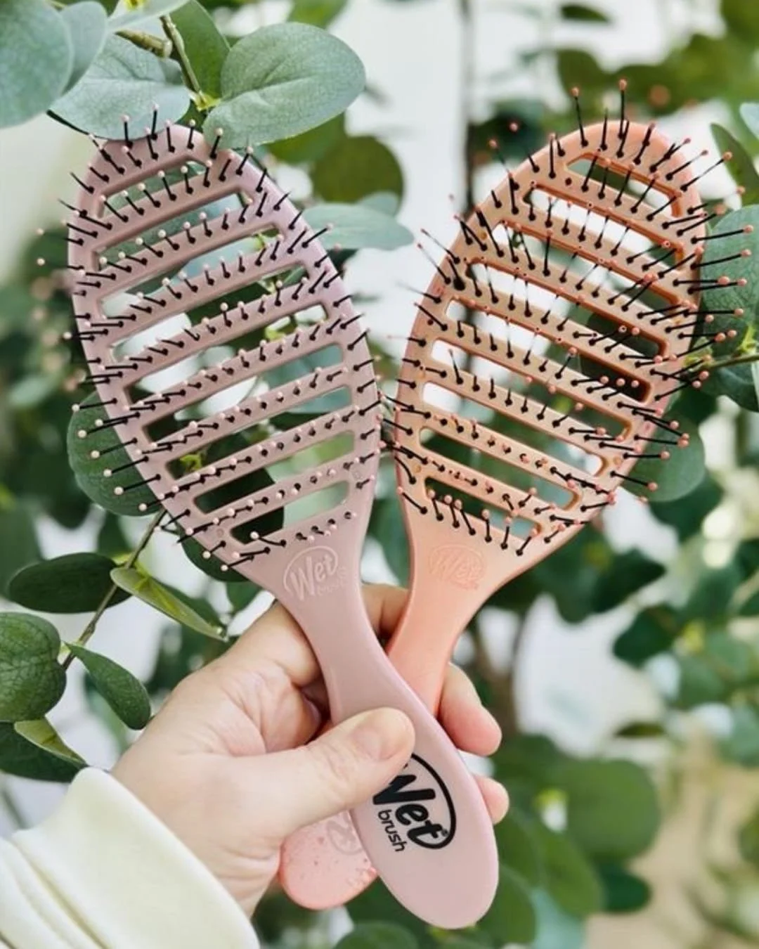Two Wet Brush Speed Dry hairbrushes in mauve and rose gold, featuring vented heads and flexible bristles, held in a hand against a backdrop of green eucalyptus leaves.
