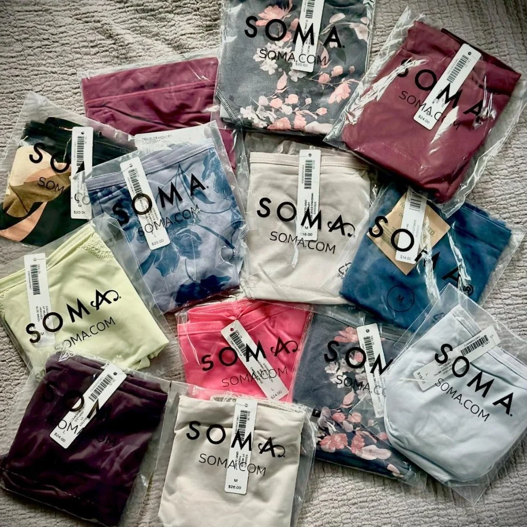 Flat lay of SOMA panties collection featuring various colors and patterns, including floral and solid designs, displayed in clear packaging with visible price tags on a textured fabric background.