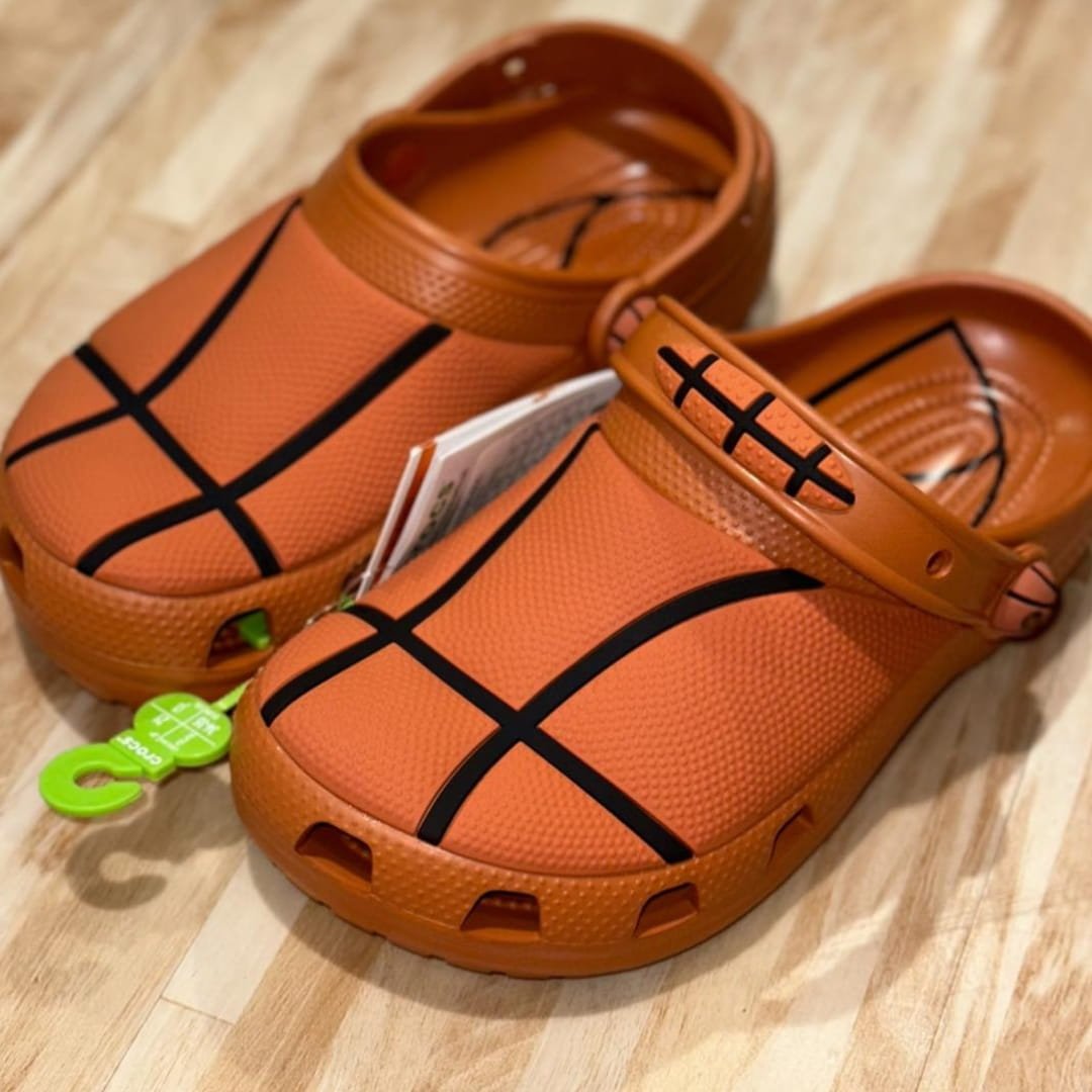 Orange Crocs sports clogs with a basketball-themed design, featuring black seam lines and a textured surface, placed on a light wooden floor with product tags attached.
