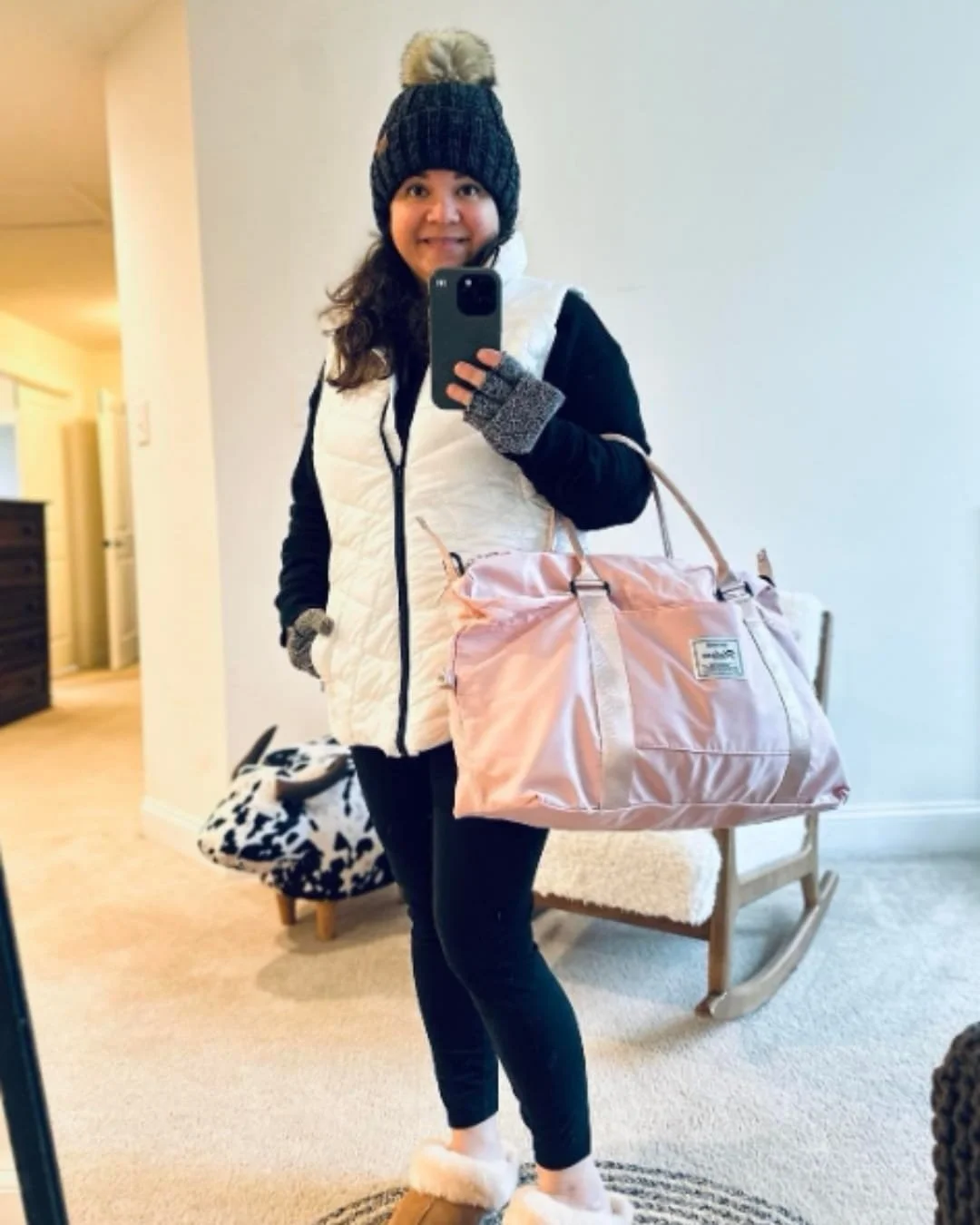 Nicole taking a selfie in cozy winter attire, wearing a beanie, quilted vest, leggings, and fuzzy slippers, holding a pink duffel bag in a warmly lit home setting.