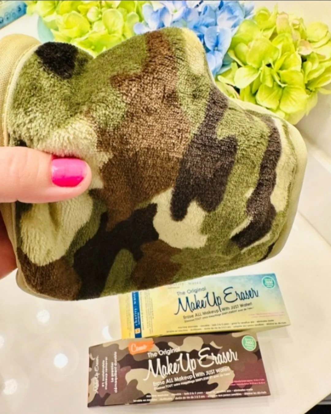 Camouflage MakeUp Eraser PRO microfiber cloth held above its product packaging on a white surface with colorful flowers in the background