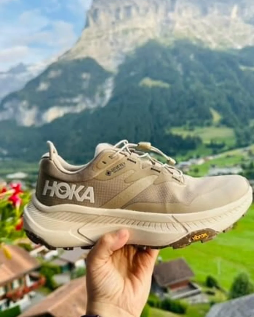 Tan Hoka hiking shoe with GORE-TEX waterproofing and Vibram outsole, held by a hand against a blurred mountain landscape featuring rocky peaks, green forests, and a lush valley.