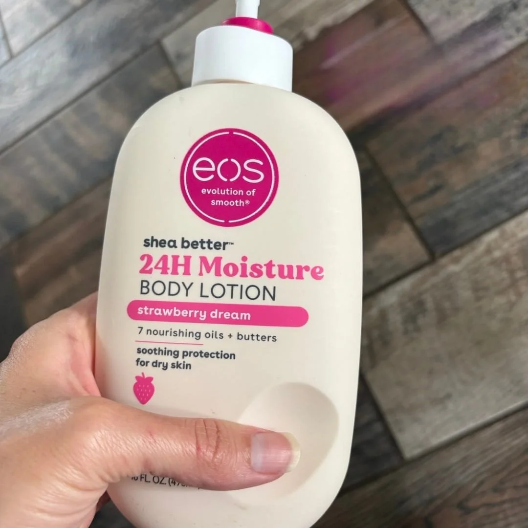 Eos Shea Better 24H Moisture Body Lotion in Strawberry Dream scent, featuring a white ergonomic bottle with a pink pump and branding, held over a wooden floor background.
