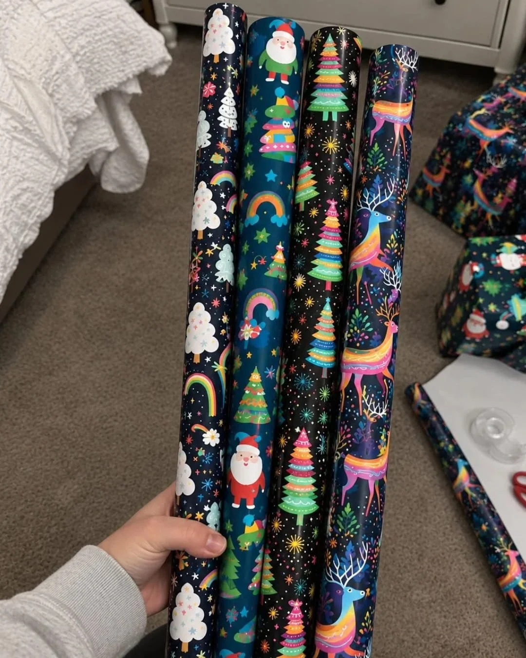 Hand holding four rolls of colorful Christmas wrapping paper with festive patterns in a cozy gift wrapping setup