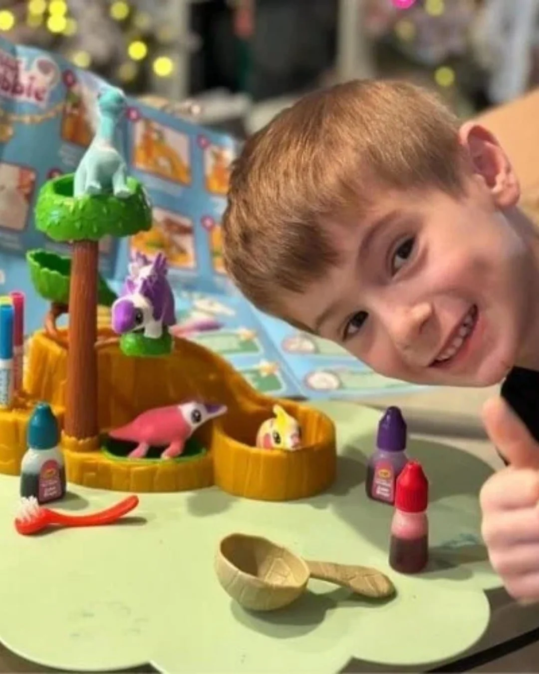 Smiling young boy giving a thumbs-up with a colorful Scrubbie toy set featuring animal figures, markers, and cleaning tools on a green mat, with a playful backdrop in a home setting.