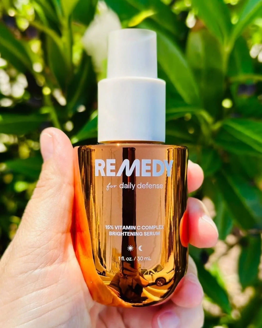 Bottle of Remedy Brightening Serum with vitamin C complex and white pump cap held outdoors against green foliage