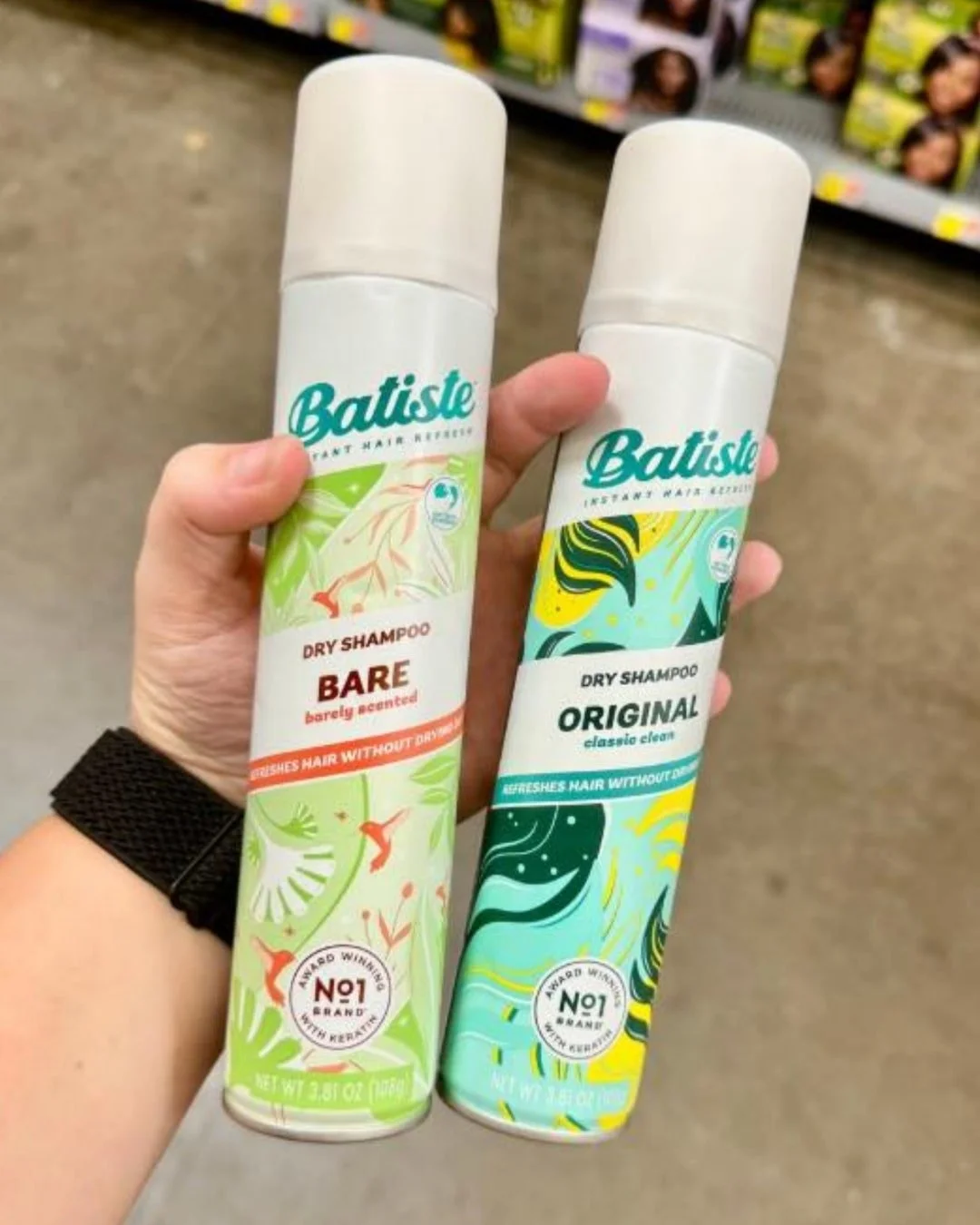 Two cans of Batiste dry shampoo, Barely Scented with a light green label and 'Original Classic Clean' with a blue label, held by a hand against a blurred retail background.