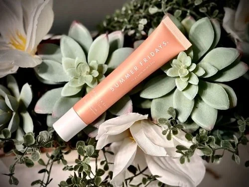 Peach-colored Summer Fridays Lip Butter Balm tube with white cap, placed diagonally over a lush background of green succulents and white flowers.