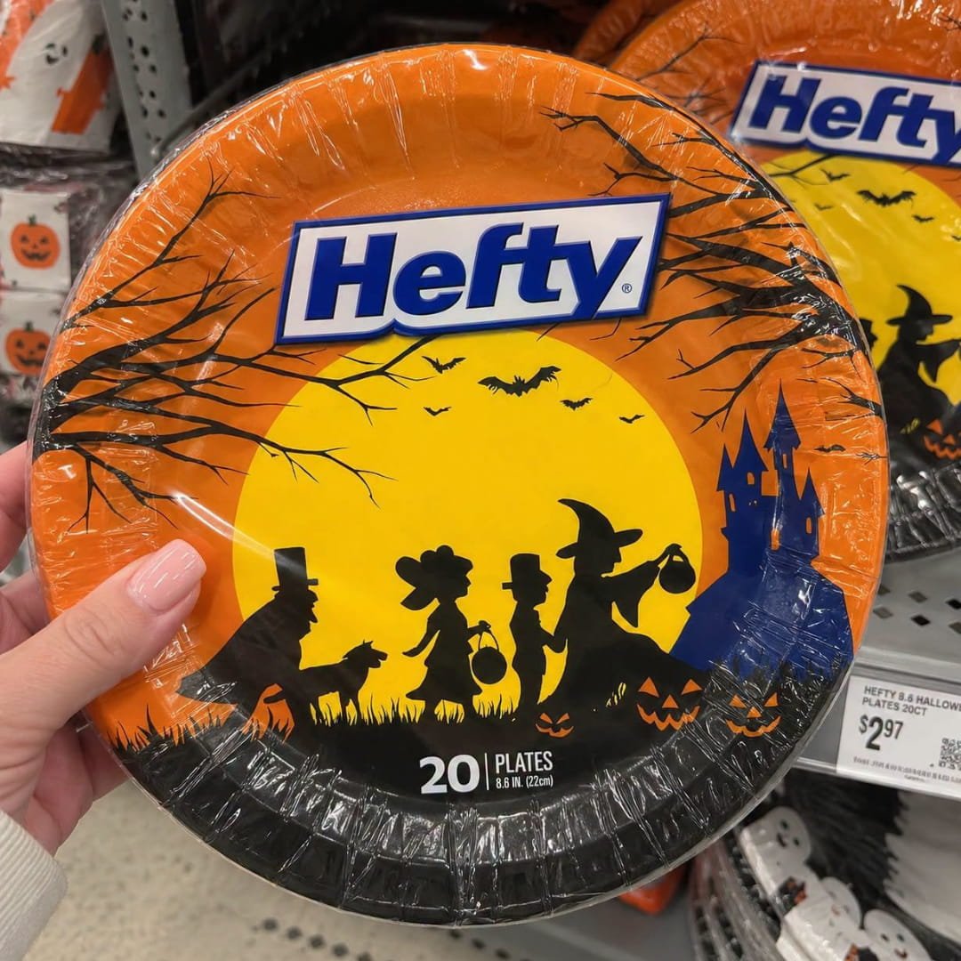 Hand holding a pack of Halloween paper plates with bats, trick-or-treaters, pumpkins, and a haunted house design