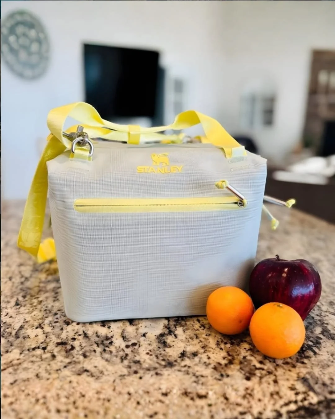 Gray Stanley insulated cooler bag with yellow accents, featuring a winged lion logo and yellow straps, placed on a granite countertop with two oranges and a red apple, suggesting its use as a food carrier in a home setting.