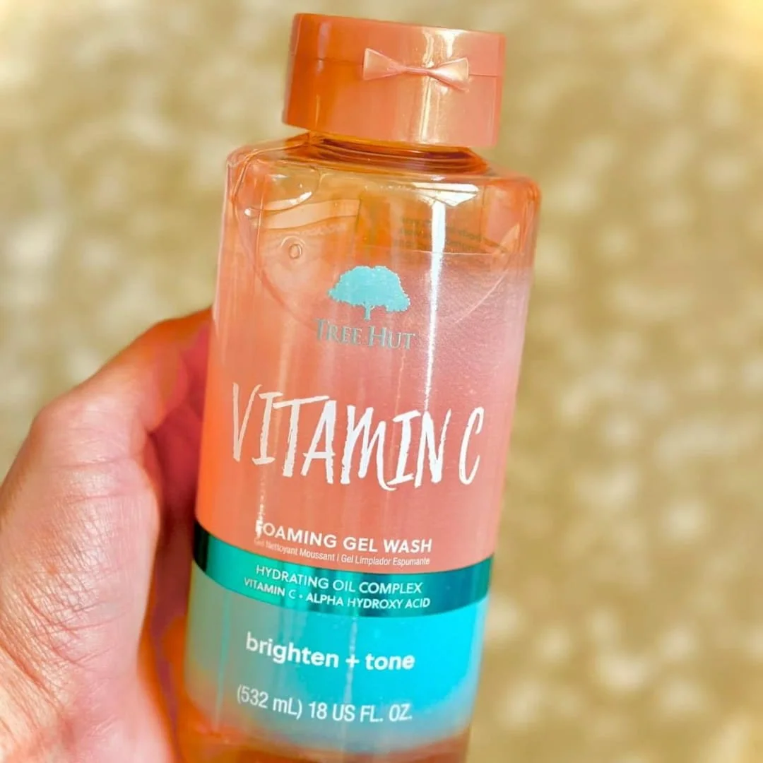 Tree Hut Vitamin C Foaming Gel Wash bottle with peachy-orange liquid, featuring hydrating oil complex and alpha hydroxy acid, held against a light textured background.