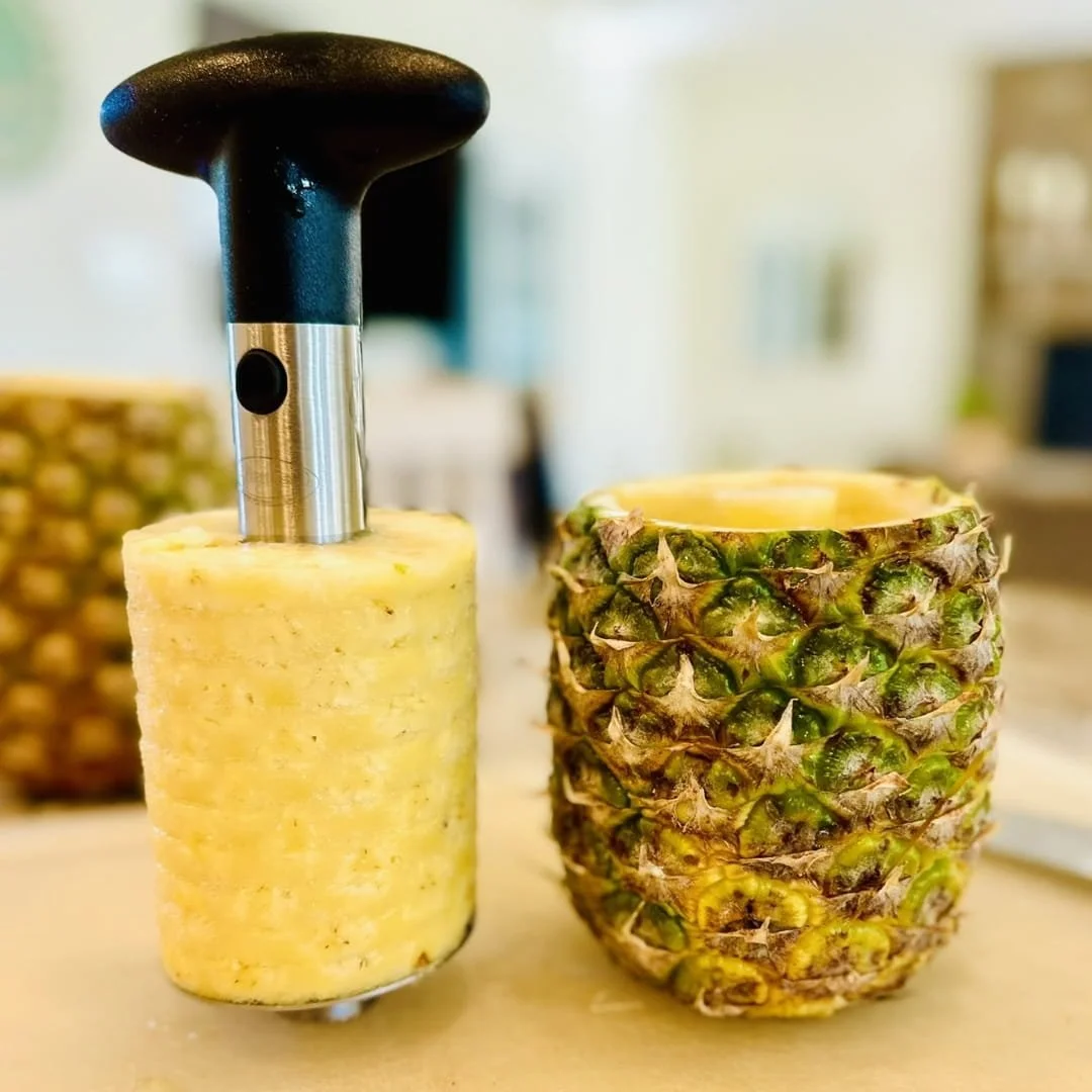 Pineapple corer and slicer tool with spiral-cut pineapple flesh beside the hollow pineapple shell on a kitchen counter