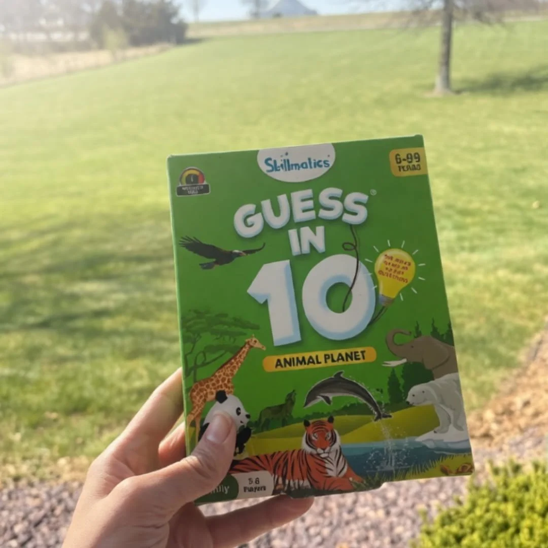 Skillmatics Guess in 10: Animal Planet game box held in a hand, featuring vibrant animal illustrations like a tiger, giraffe, and dolphin, with a green background and outdoor grassy setting.