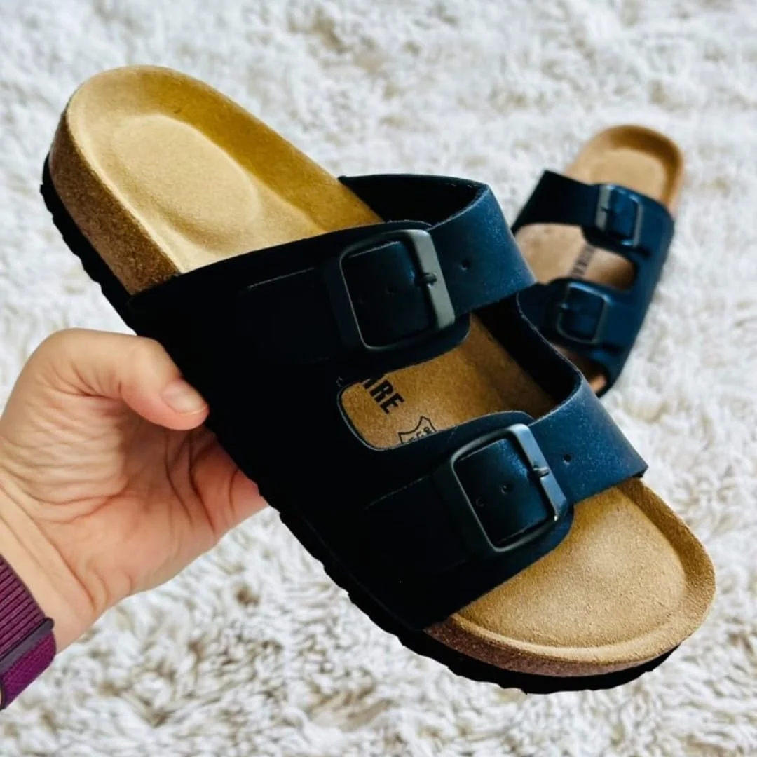 CUSHIONAIRE Luna sandals with black double straps, adjustable buckles, a tan suede footbed, and cork-textured midsole, displayed on a white shaggy rug with one sandal held in hand.