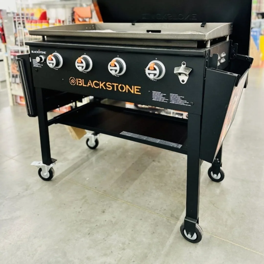 Blackstone 4-burner outdoor gas griddle with open hood, flat cooking surface, four control knobs with orange accents, built-in bottle opener, and side storage bins, displayed in a retail store.