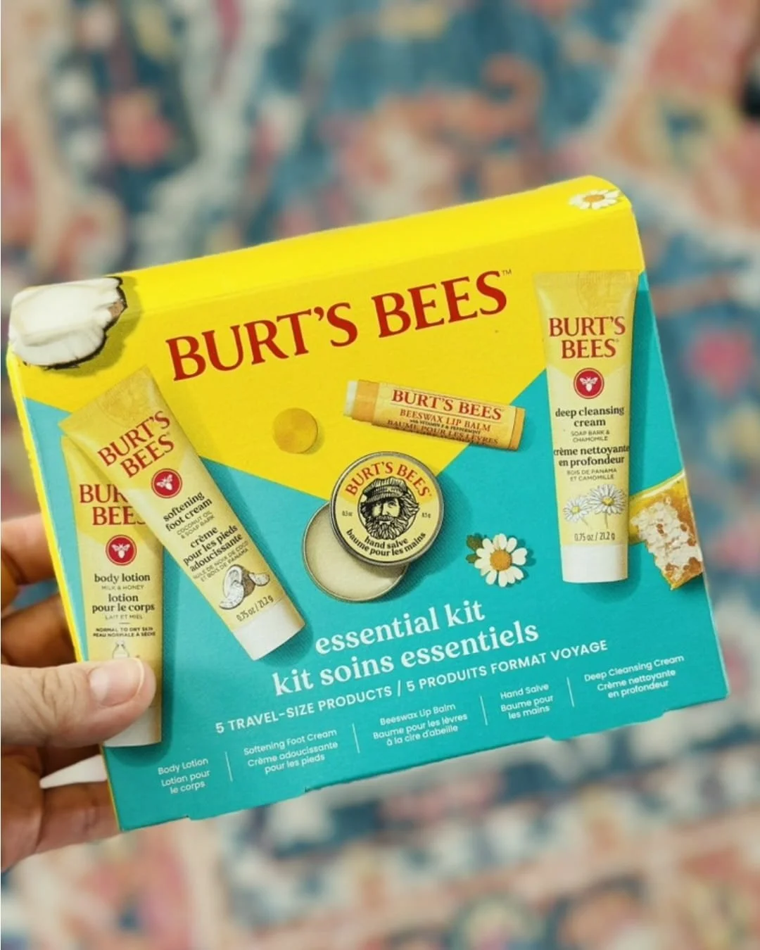 Burt's Bees Essential Kit gift box featuring five travel-sized skincare products, including body lotion, foot cream, lip balm, hand salve, and cleansing cream, with vibrant yellow and teal packaging held in a hand.