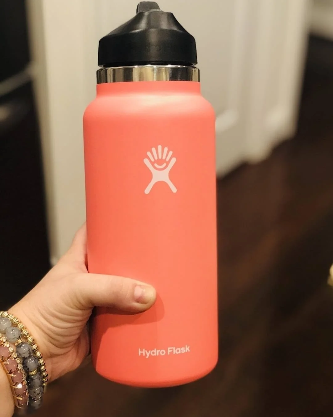 Coral-colored Hydro Flask water bottle with a matte finish, featuring a black straw lid and white logo, held by a hand adorned with beaded bracelets against a softly blurred indoor background.